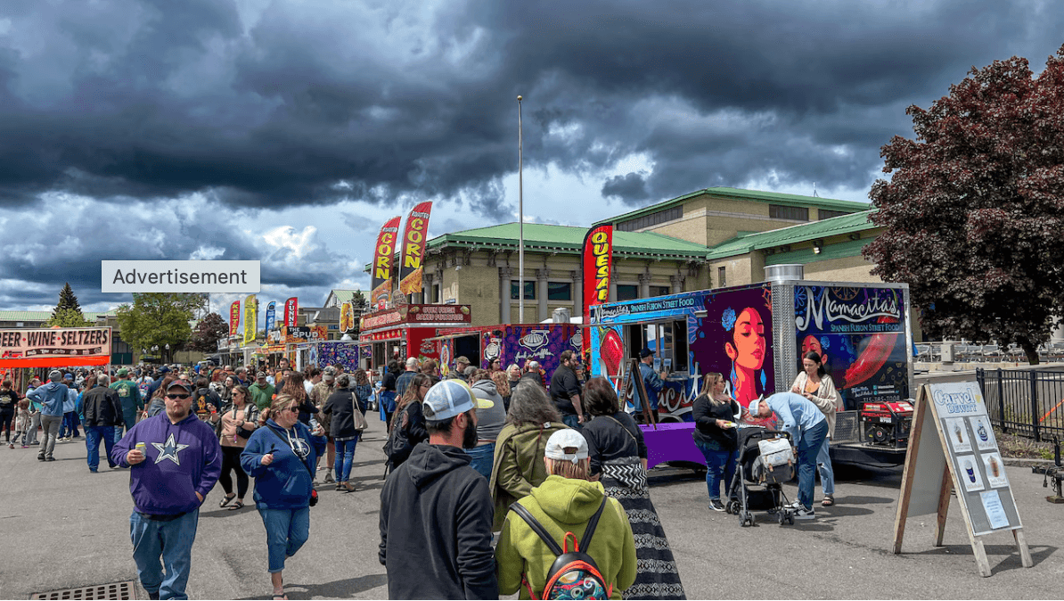 Syracuse Food Truck Battle New York State Fairgrounds NYS Crowds walk past colorful food trucks and stalls at an outdoor fair under a dramatic, cloudy sky. At events like Crafts at Lyndhurst, flags advertising corn and quesadillas wave as people enjoy eating and socializing with friends.